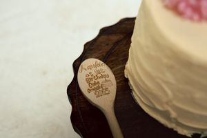 Close-up of an engraved wooden spoon beside a white frosted birthday cake on a dark wood stand.