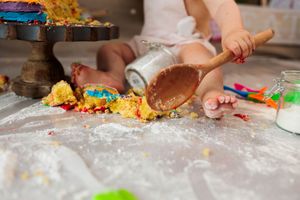 Baby smashing colorful rainbow cake on flour-covered surface with wooden spoon during photoshoot.