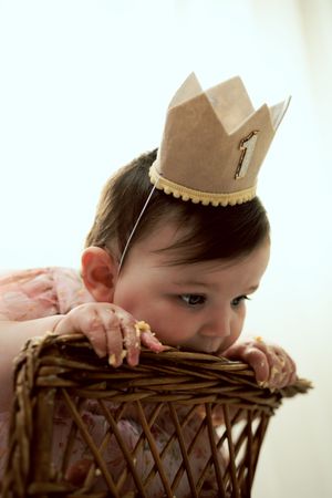 Baby girl wearing a paper crown peering over a wicker basket at her first birthday cake smash.