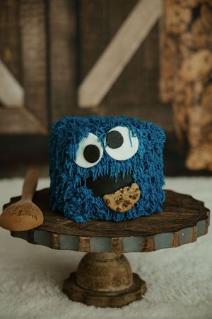 Blue Cookie Monster character cake with googly eyes and chocolate chip cookie on a rustic wooden cake stand.