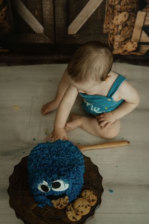 Baby in blue overalls reaching toward a Cookie Monster themed first birthday cake smash on a wooden floor.