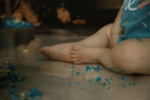 Baby's chubby feet and legs sitting on floor with blue cake crumbs during first birthday smash cake celebration.