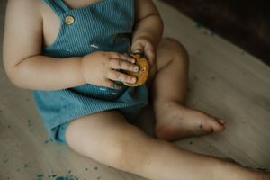 Baby in blue romper sitting on floor eating a cookie with chubby hands and bare feet.
