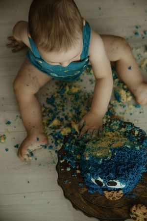 Baby smashing blue frosted first birthday cake, wearing teal suspenders, with cake crumbles scattered on white floor.
