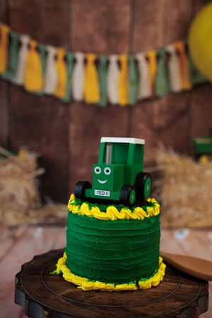 Green tractor-themed first birthday smash cake with yellow frosting on rustic wooden stand.