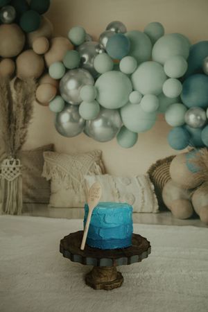 Blue ombre smash cake on wooden stand with teal and silver balloon garland backdrop and boho pillows.
