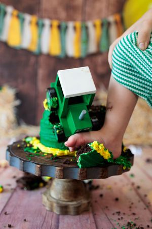 Baby hand smashing green tractor birthday cake on wooden cake stand during cake smash session.