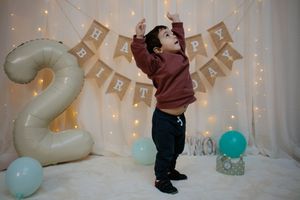 Toddler celebrating second birthday with arms raised beside a number 2 balloon and colorful decor.