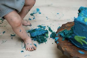 Baby feet standing next to smashed blue birthday cake pieces on white floor.