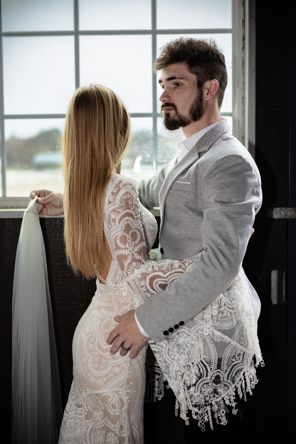 Couple in wedding attire embraces near window in intimate portrait.