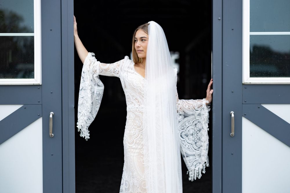 Bride in white lace gown poses elegantly between modern doors with flowing veil.