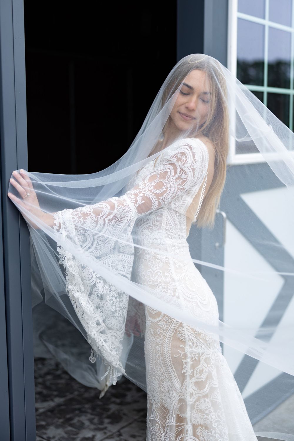 Bride in lace wedding dress twirls her veil in sunlit doorway.