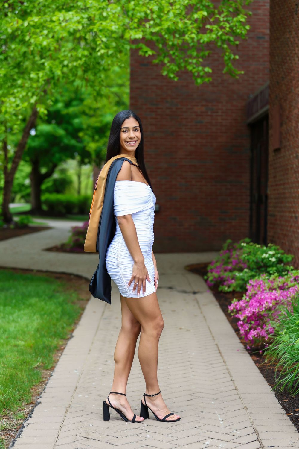 A smiling graduate in a white dress poses with her graduation gown on a campus walkway.
