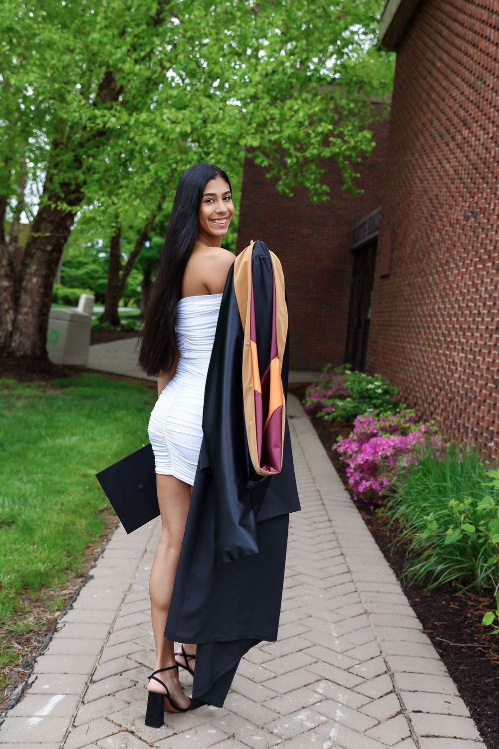 A graduation photo of a woman in a white dress and academic regalia on a brick-lined path.