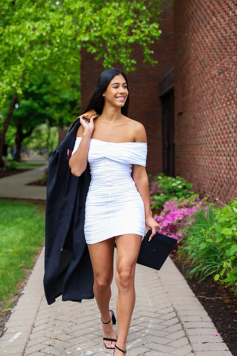 A graduate in a white off-shoulder dress carries her cap and gown while walking on campus.