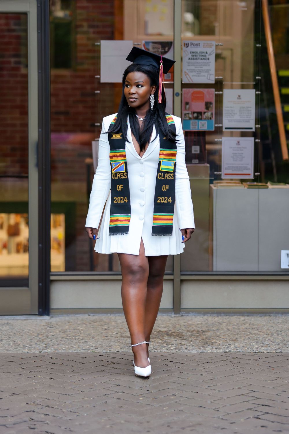 Graduate wearing white attire and decorated stole walking confidently outside campus building.