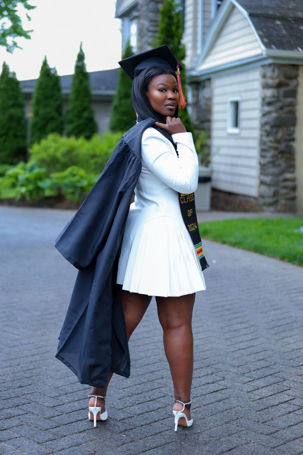 Graduate in white dress and black graduation gown posing with silver heels on brick pathway.