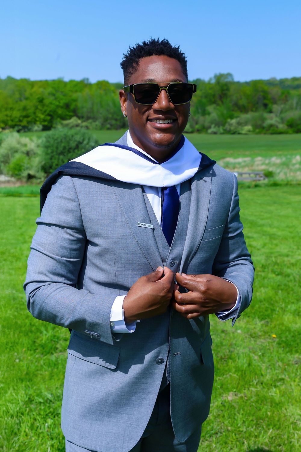 Person in grey blazer and sunglasses smiles while adjusting blue tie outdoors.