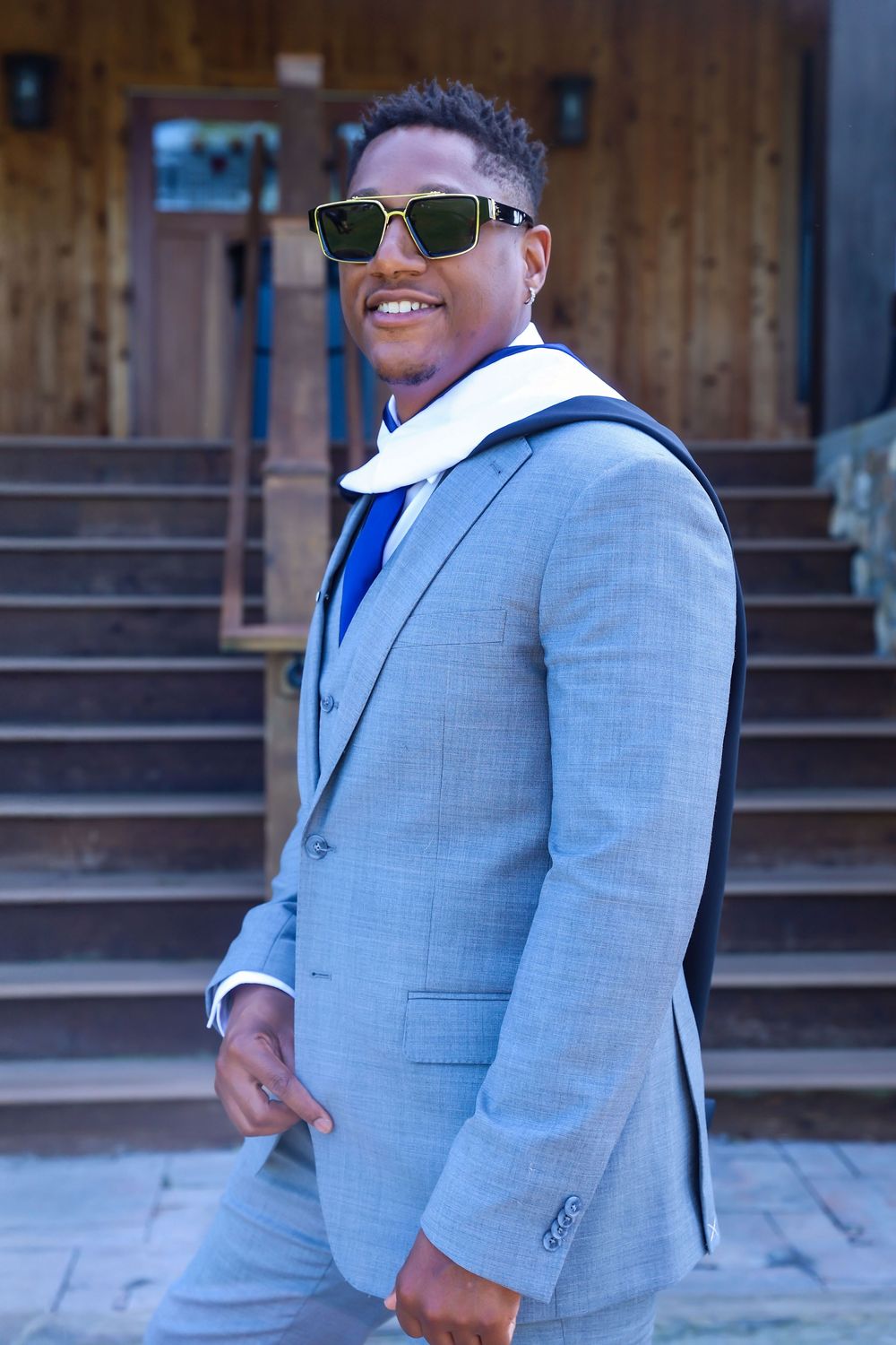 Person in grey suit poses confidently outside wooden building entrance.