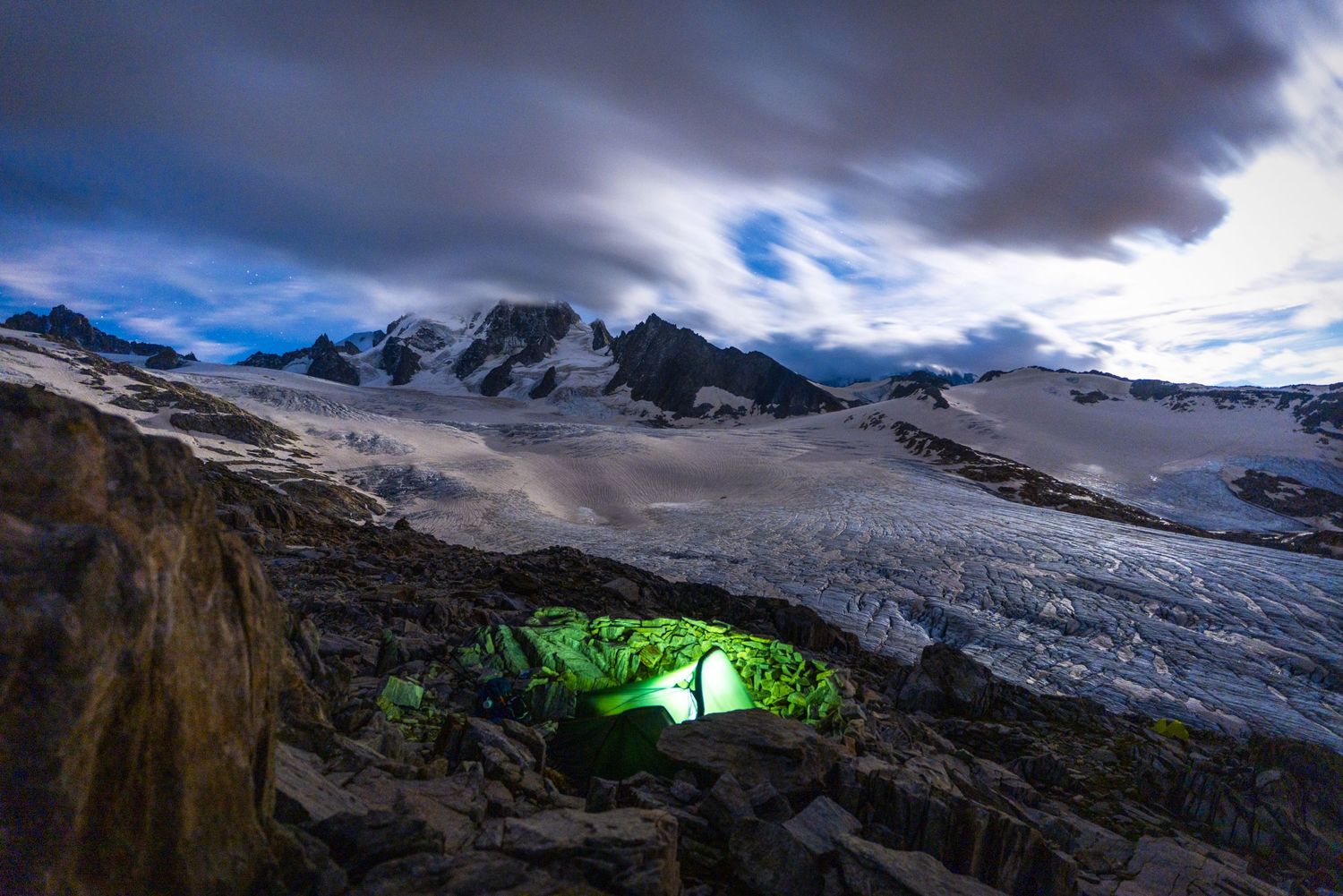 Glowing green tent illuminated beneath snowy mountain peaks and dramatic night sky.