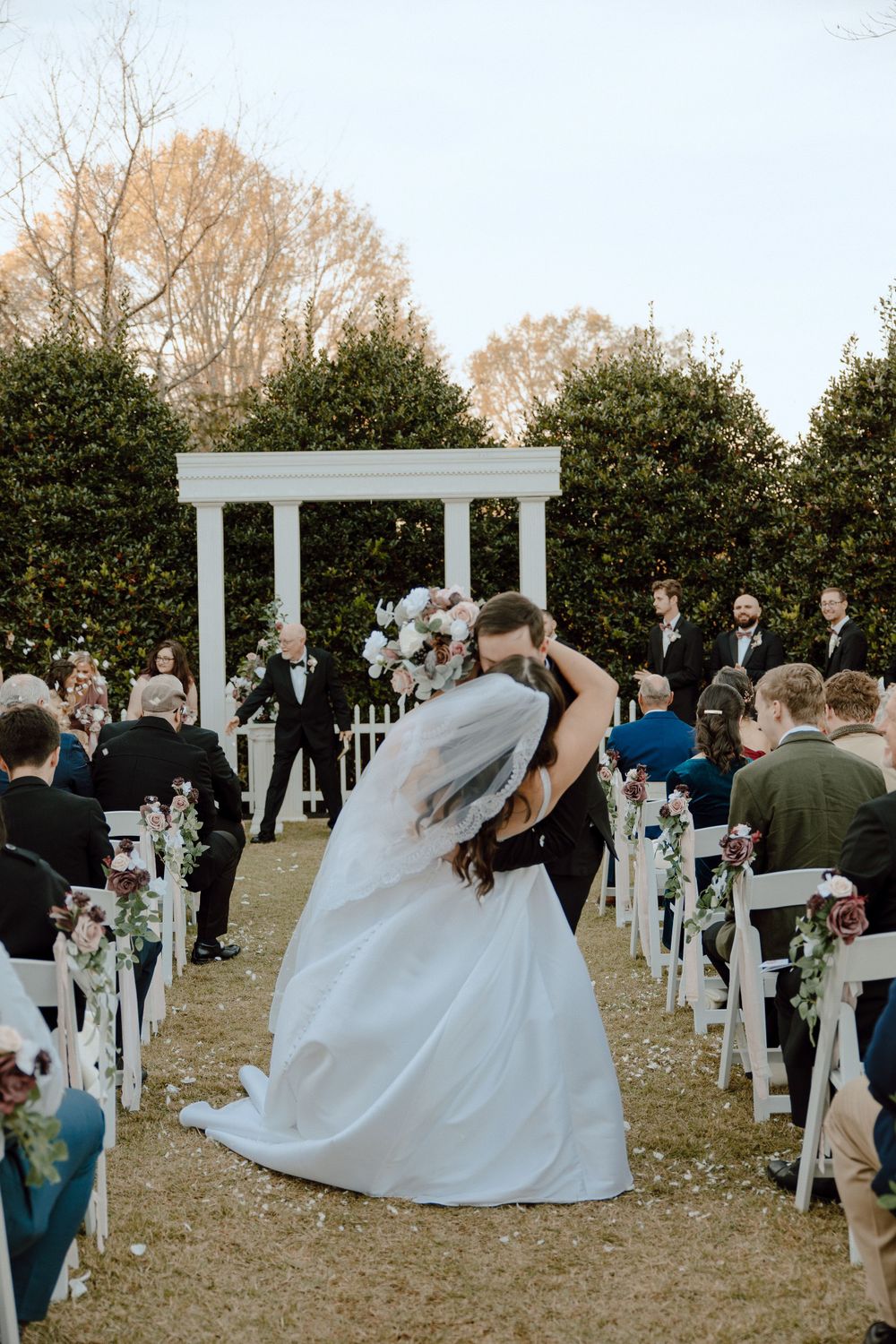A wedding couple sharing their first kiss at an outdoor ceremony with white chairs and decorations.