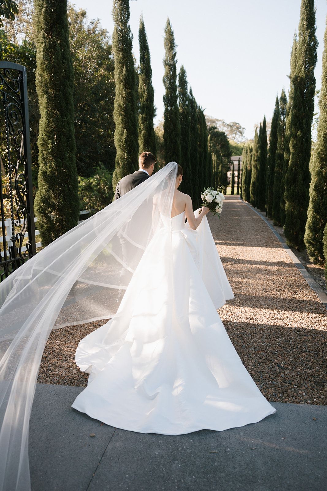 A couple in elegant wedding attire walks together on grass during golden hour.