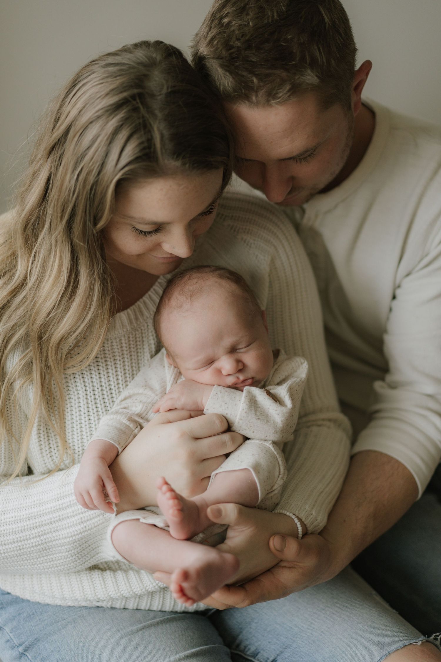 Loving parents in neutral tones cuddle sleeping newborn baby together indoors.