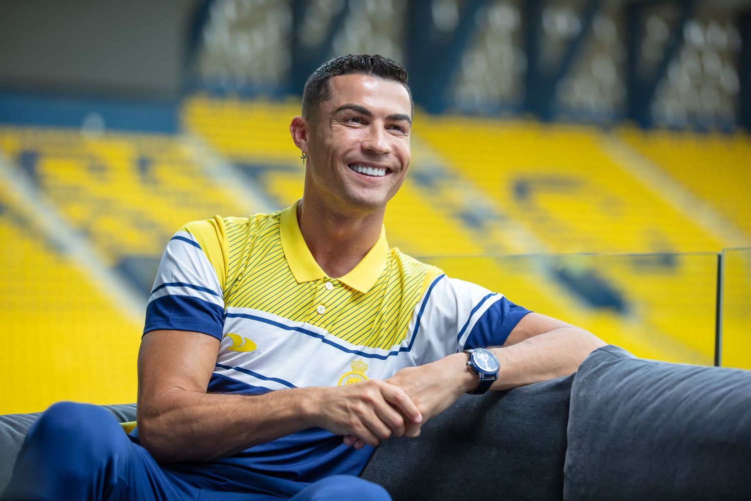 Soccer player in yellow training vest sits smiling in stadium with yellow seats in background.
