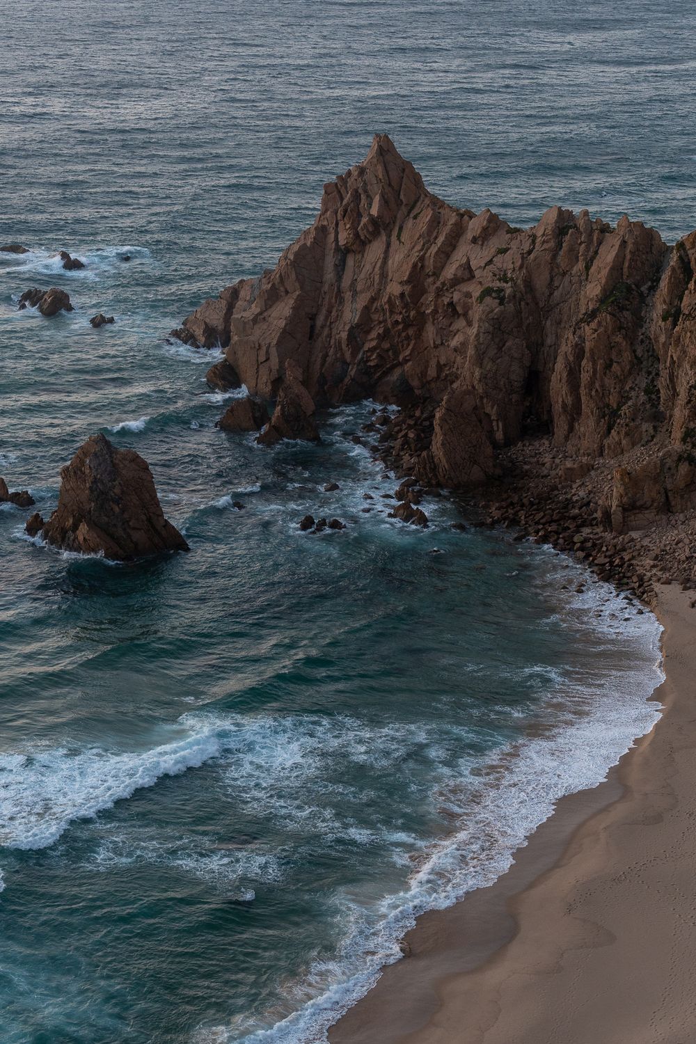 Dramatic rocky coastal cliffs rising above a sandy beach with crashing waves.