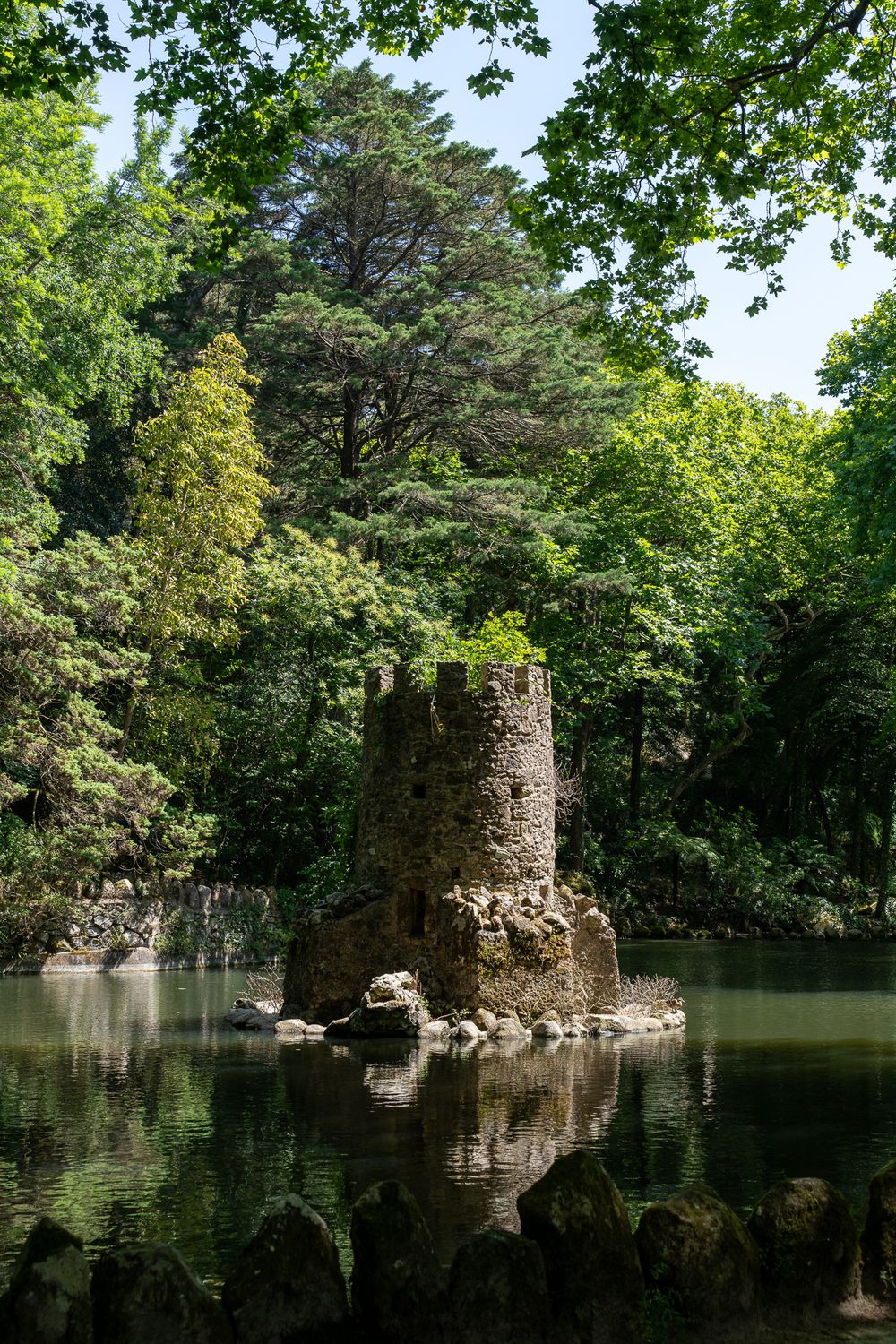 Ancient stone tower ruin reflected in a peaceful pond surrounded by lush forest.
