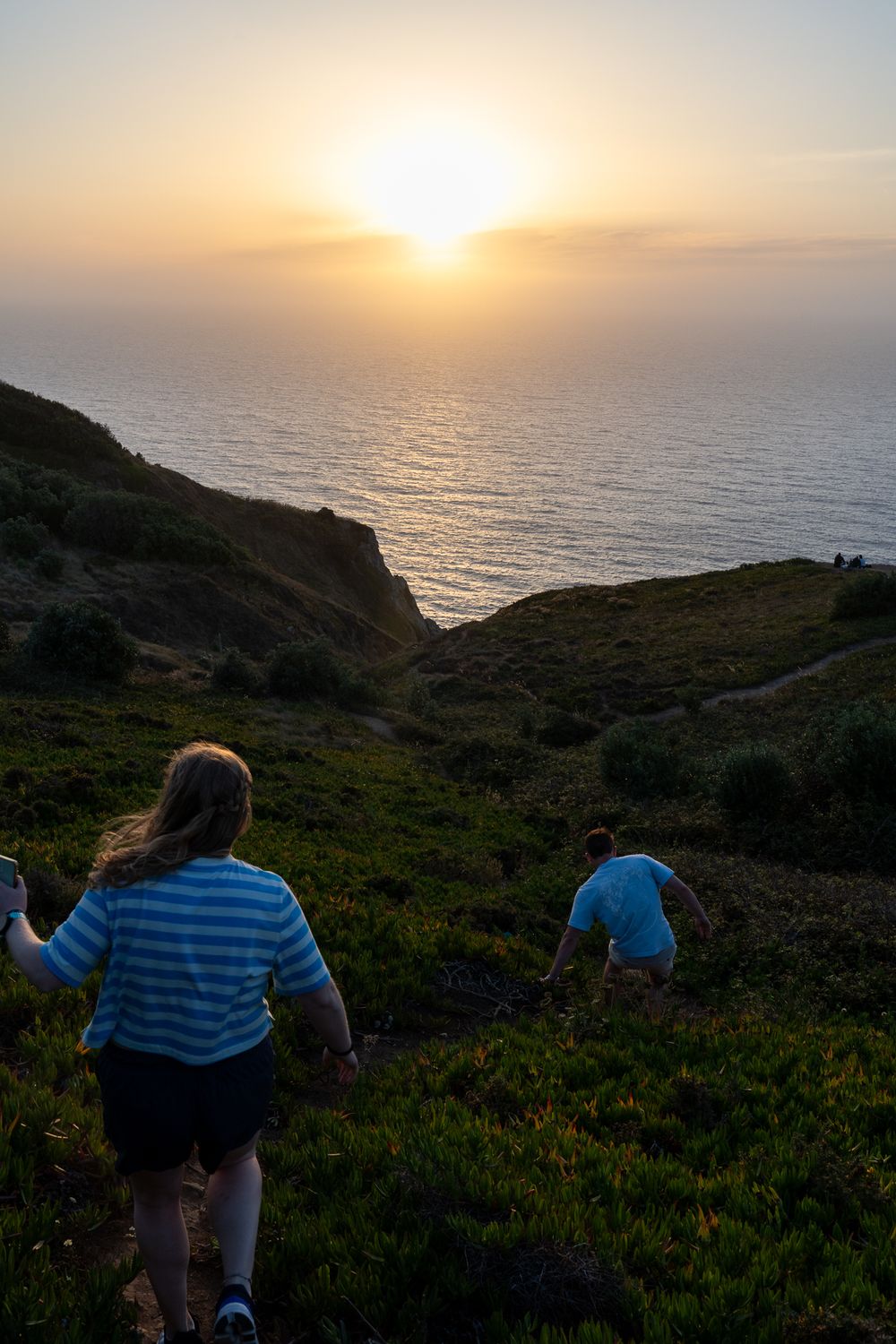 Scenic coastal sunset view with people hiking down a grassy hillside path.