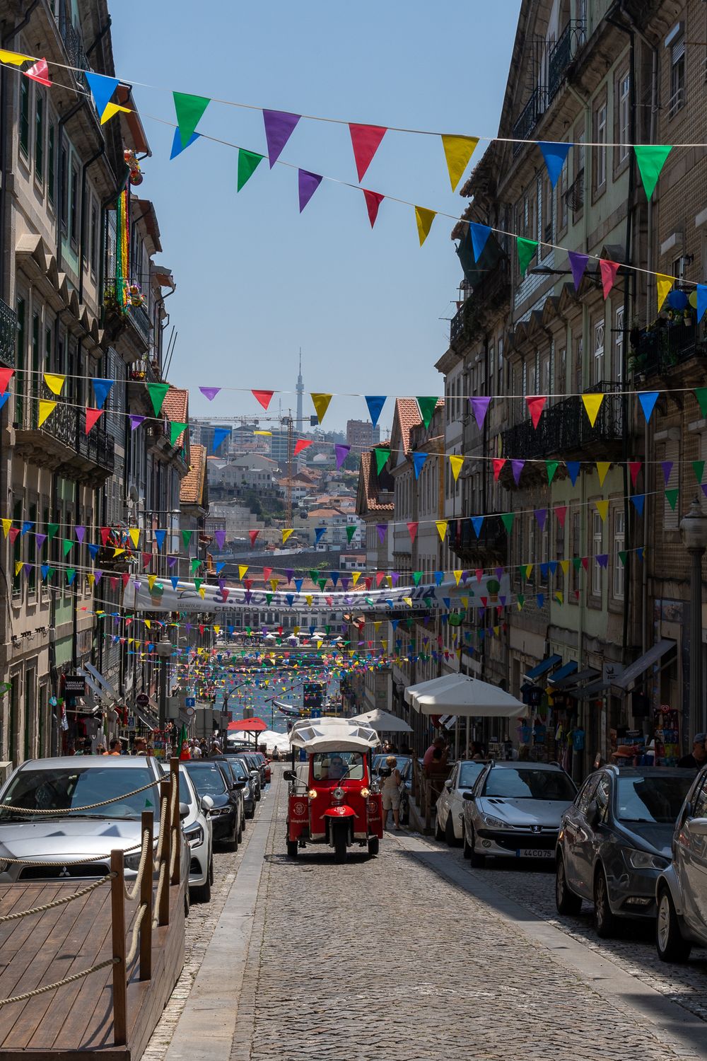 Colorful triangle flags hang across a narrow cobblestone street with parked cars and historic buildings in Portugal.