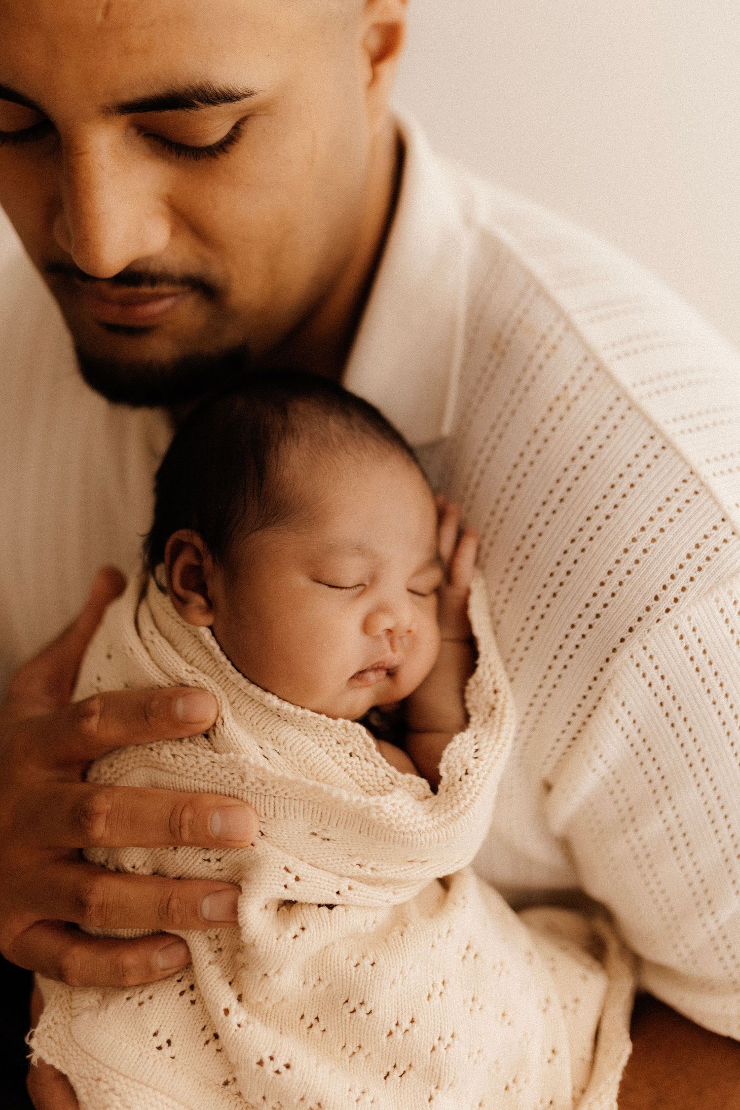 Tender moment between parent and sleeping newborn wrapped in white knit blanket.