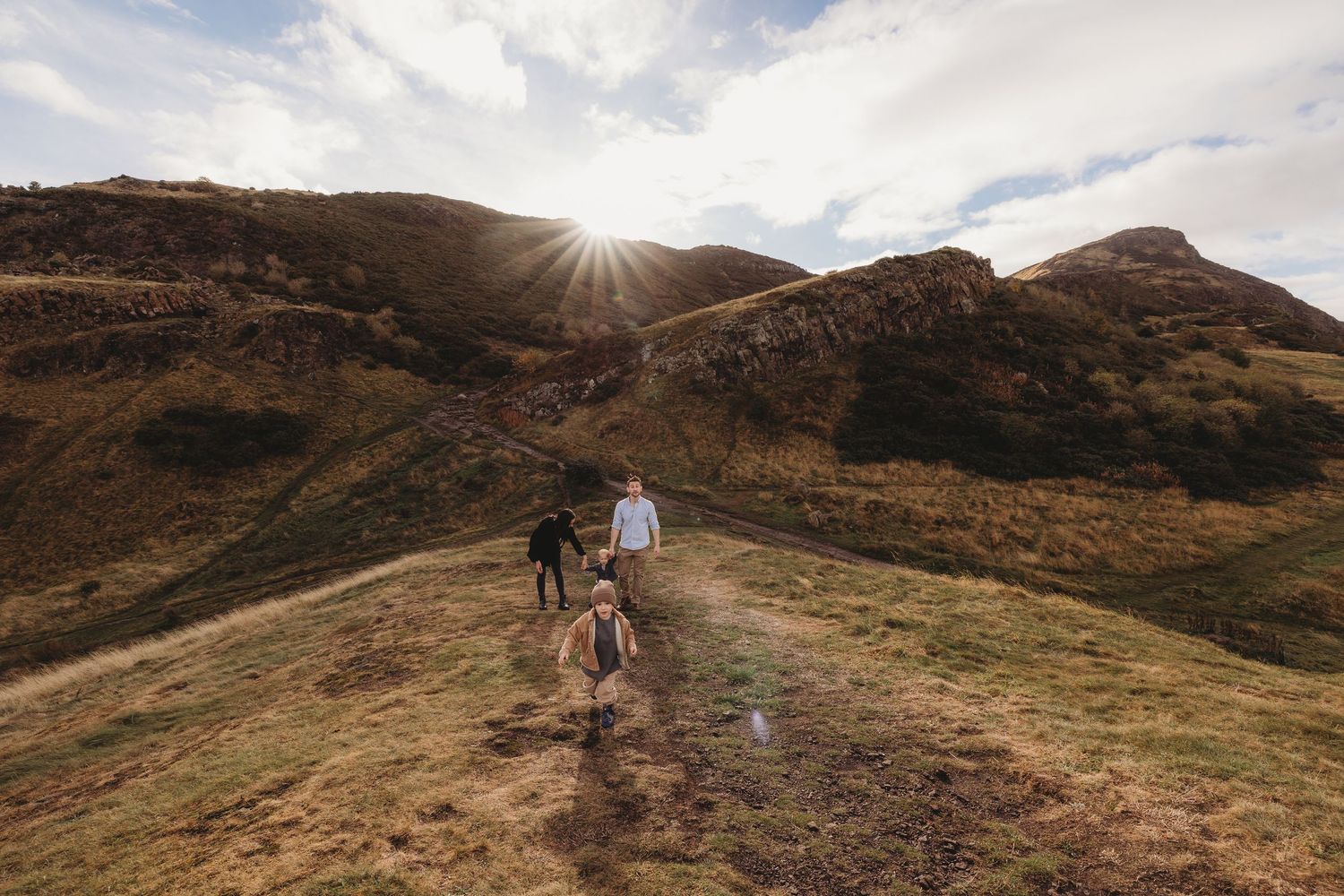 Two hikers walk along a scenic mountain trail during golden hour with dramatic clouds and rugged terrain in view.