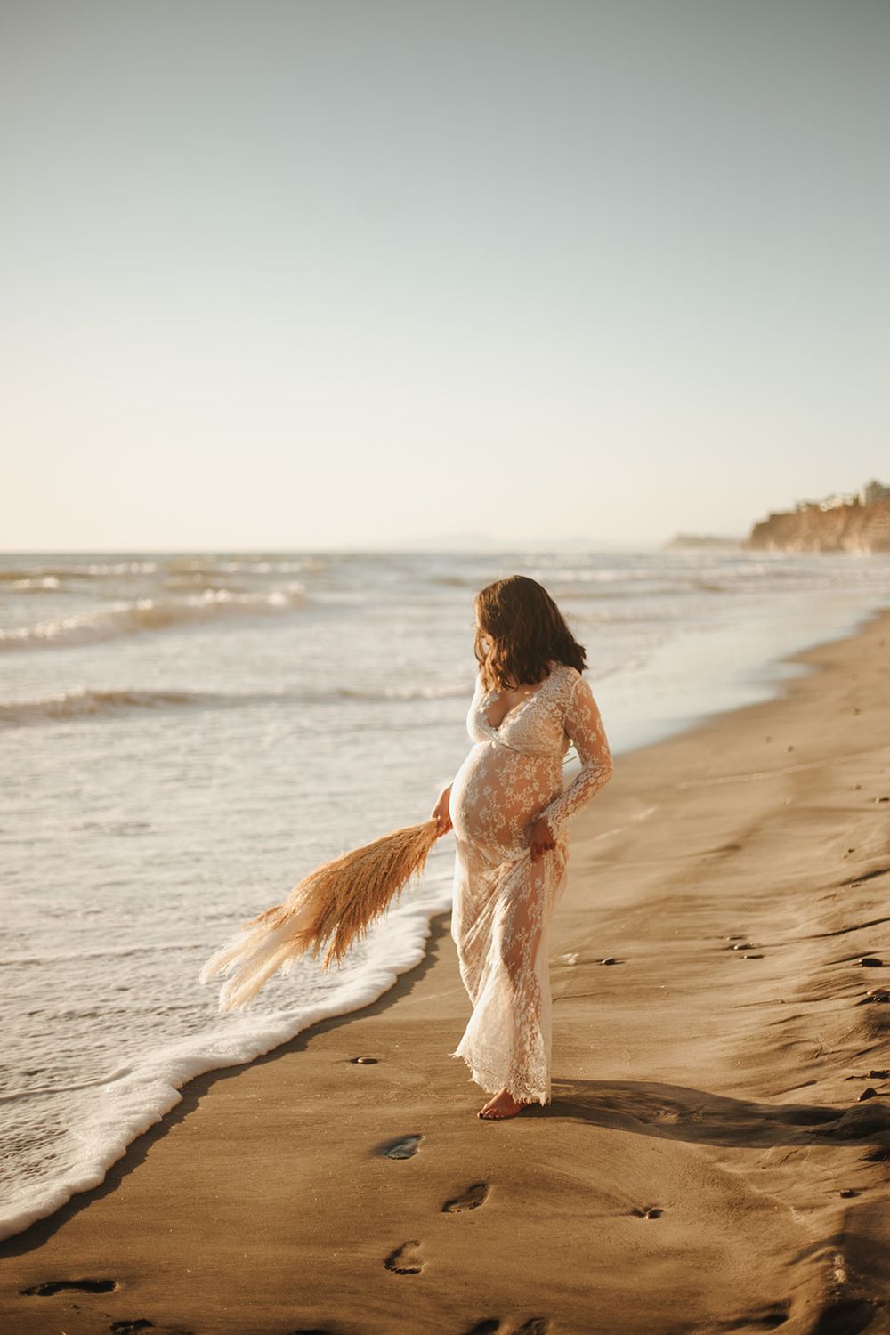 Pregnant woman standing on the beach, gazing at the ocean with the sun setting in the background.