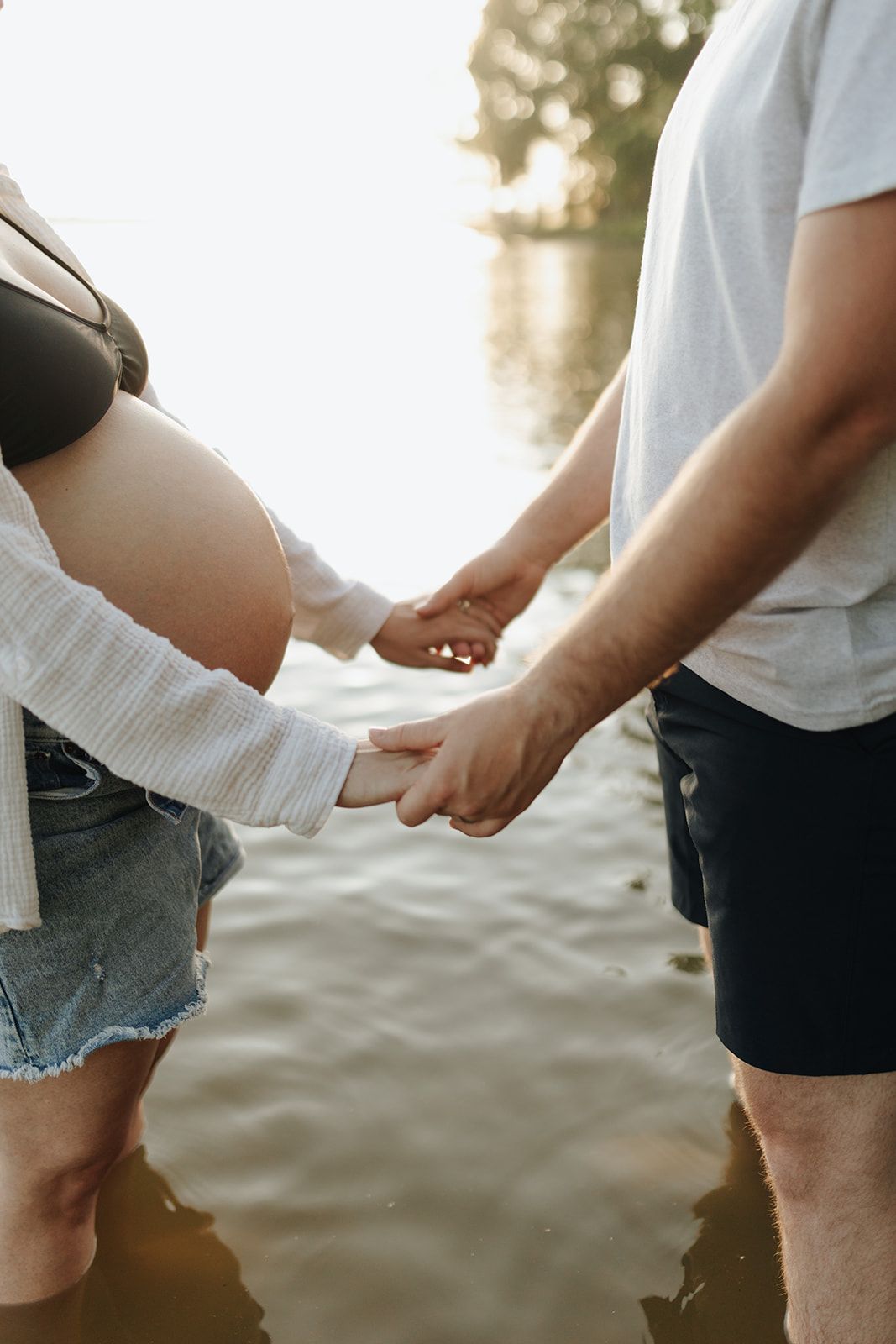 Close-up shot of a pregnant couple wading in a lake, with the woman's baby bump visible as they stand hand-in-hand.