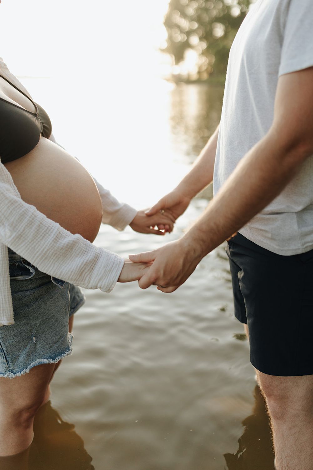 Close-up shot of a pregnant couple wading in a lake, with the woman's baby bump visible as they stand hand-in-hand.