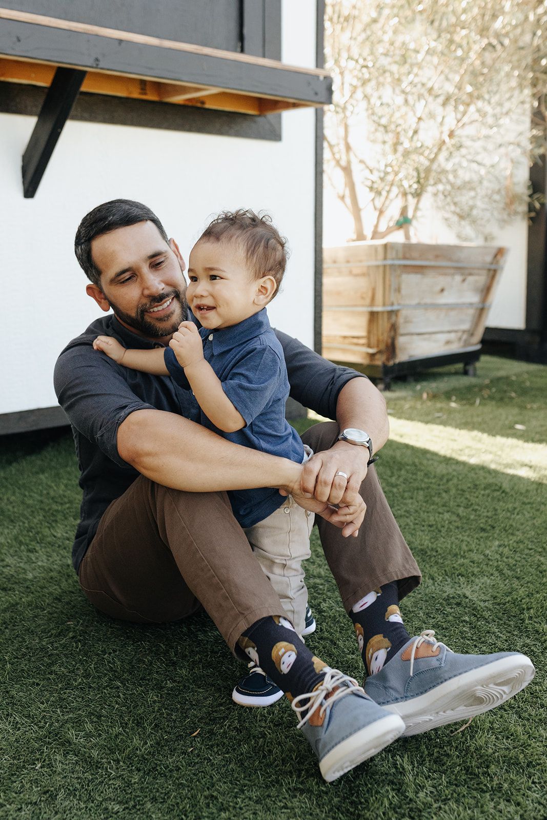 Father sitting with his young son, smiling and sharing a moment outdoors, with the father holding his son in his arms.