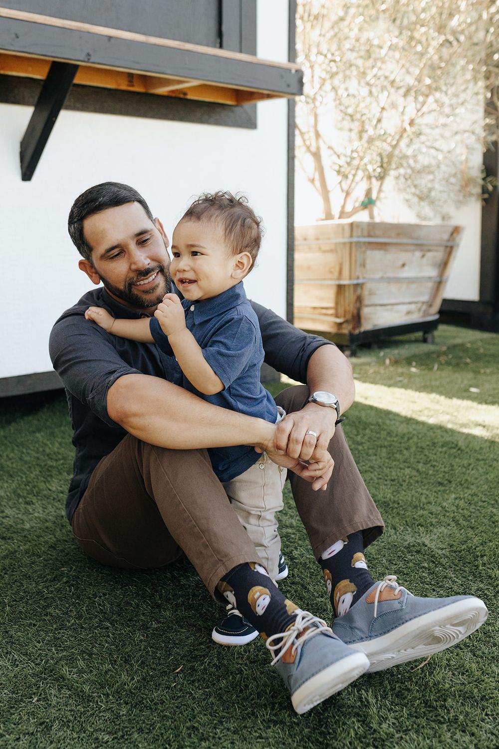 Father sitting with his young son, smiling and sharing a moment outdoors, with the father holding his son in his arms.