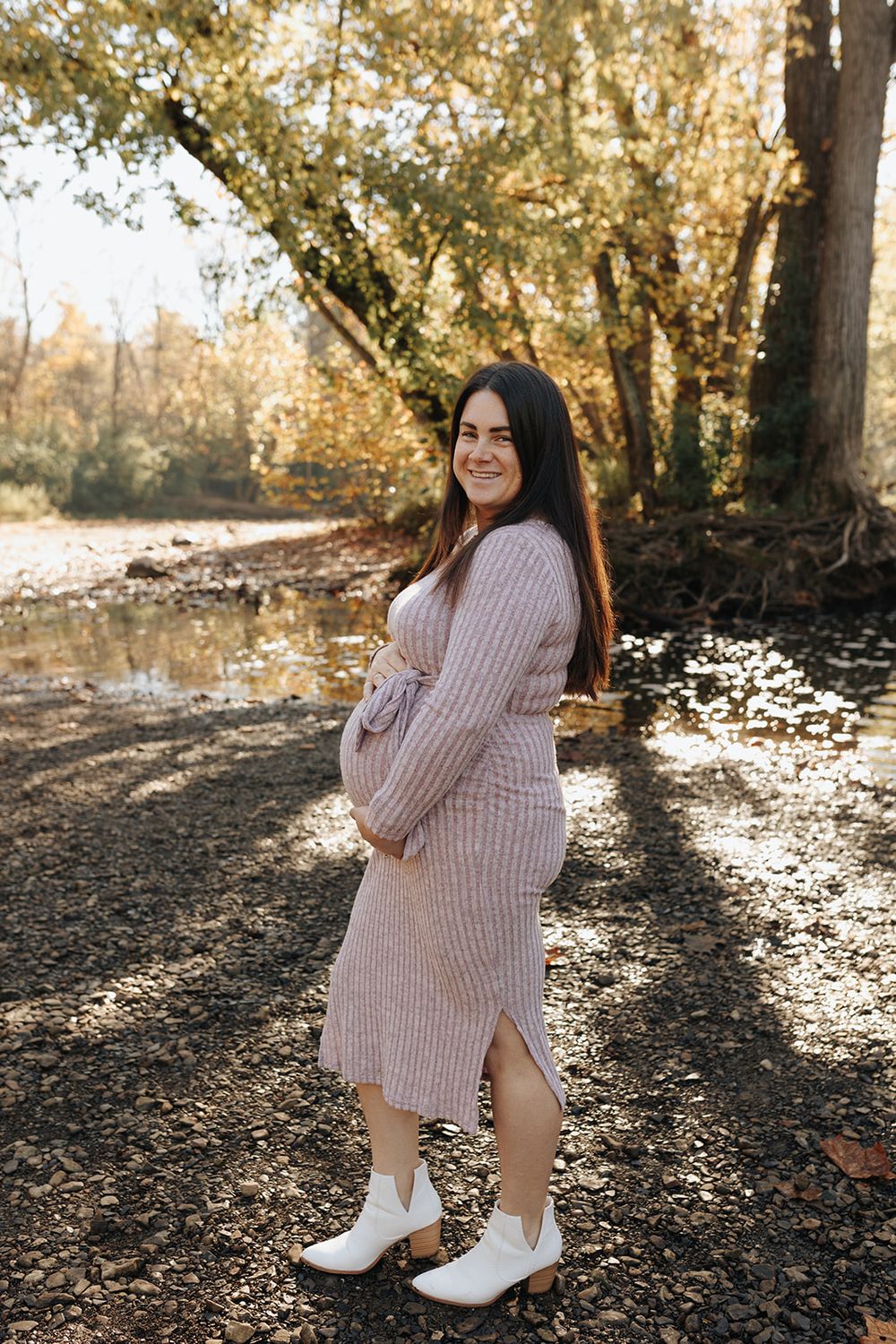 Pregnant woman standing by a river surrounded by autumn foliage, with warm golden and orange leaves in the background.