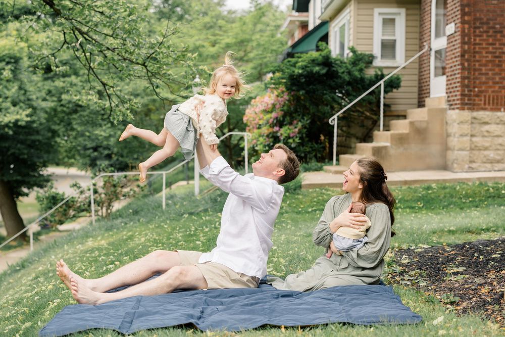Family enjoying a sunny day outdoors, father lifting toddler in air while mother relaxes on blue blanket in green yard.