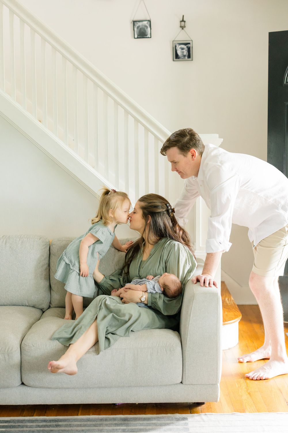 A young family shares a tender moment on a gray sofa, with mom holding a newborn while toddler kisses her cheek.