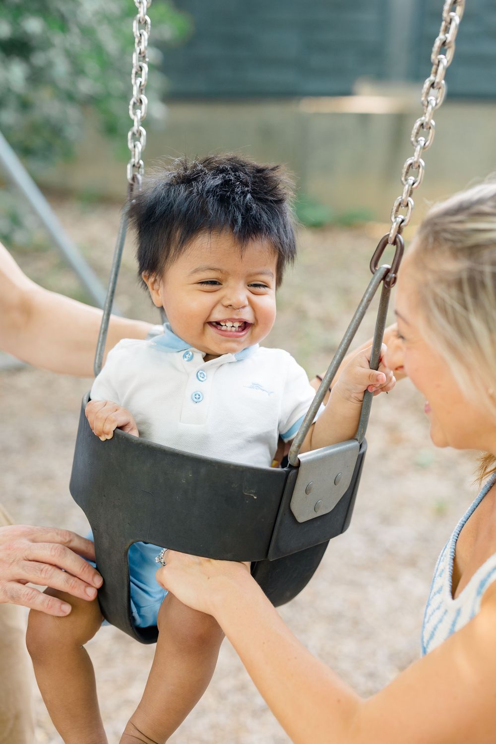 Happy baby laughing on a toddler swing being pushed by a smiling blonde woman at a playground.