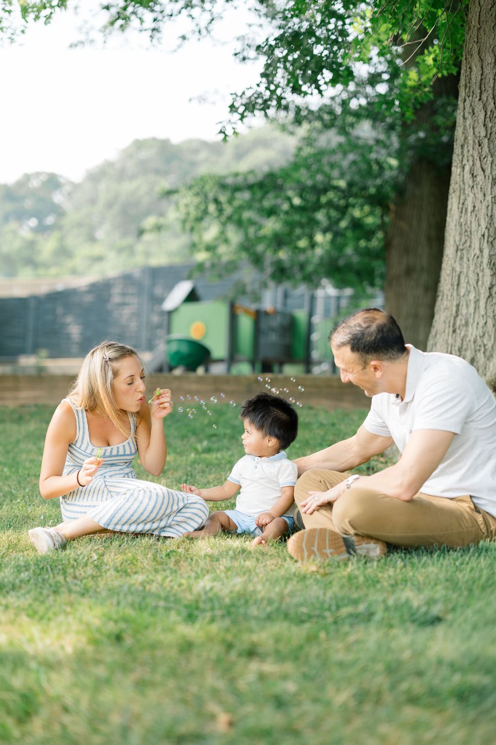 A happy family sits on green grass under a tree, with a mother blowing dandelion seeds to her toddler.