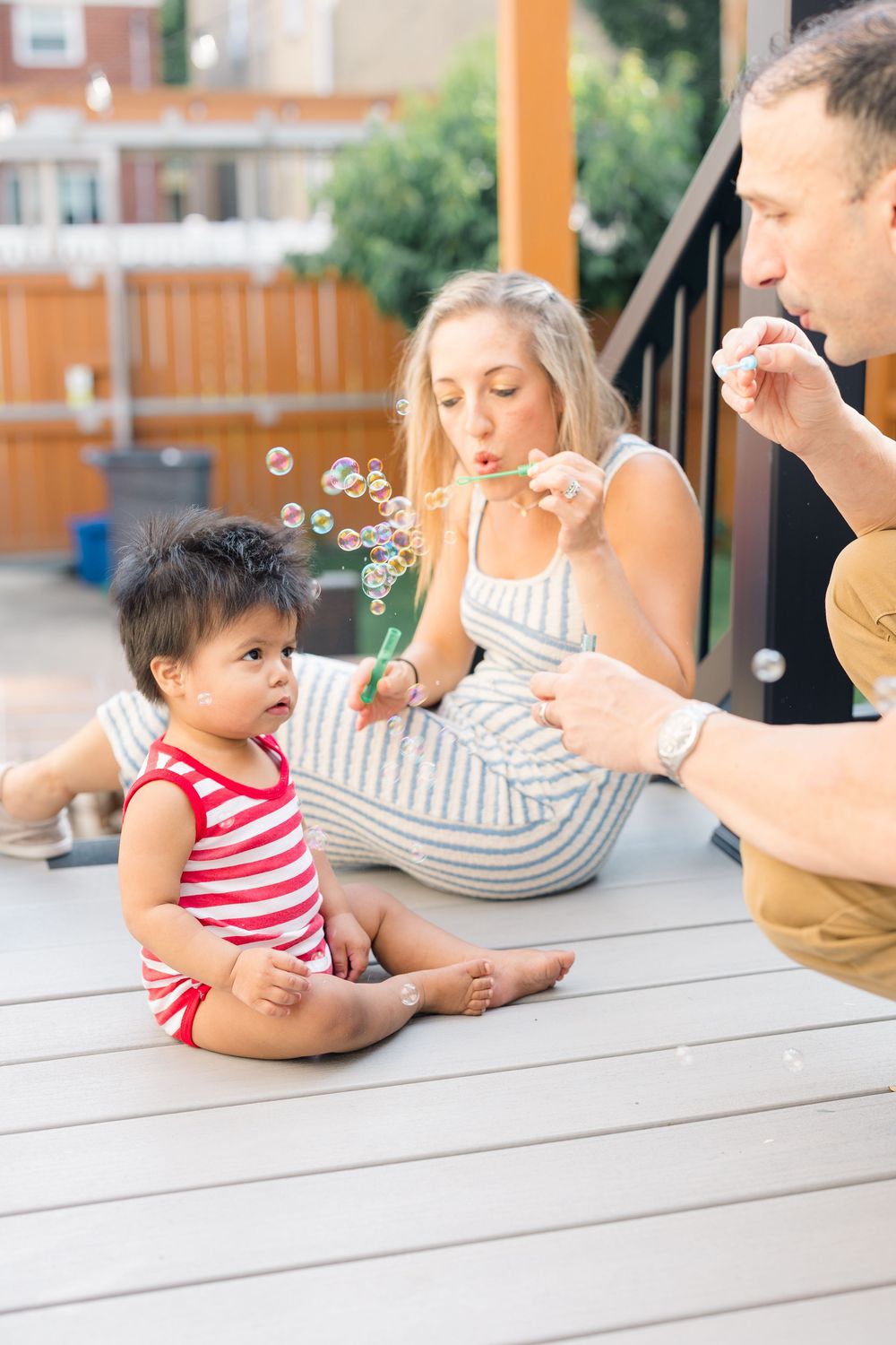 Family blowing bubbles with toddler on outdoor deck, parents entertaining baby with bubble wands on summer day.