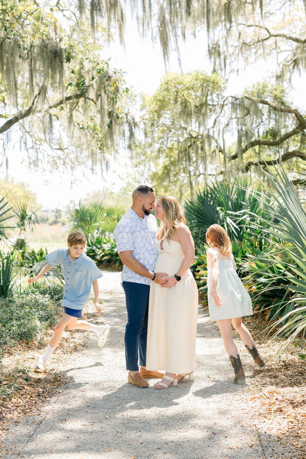 A couple shares a tender moment on a garden path while two children play around them, framed by moss-draped oak trees.