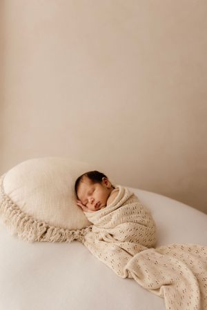 Newborn baby sleeping on beige pillow wrapped in cream knit blanket.