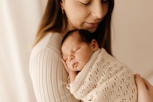 Mother in white sweater holding sleeping newborn wrapped in knit blanket.