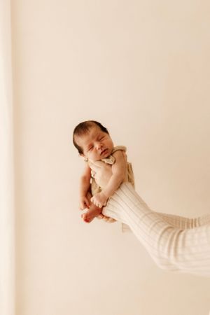 A newborn baby wearing a cream-colored outfit yawns while being held against a neutral background.