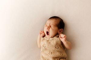 A peaceful sleeping newborn in a beige knit outfit stretches against a light backdrop.